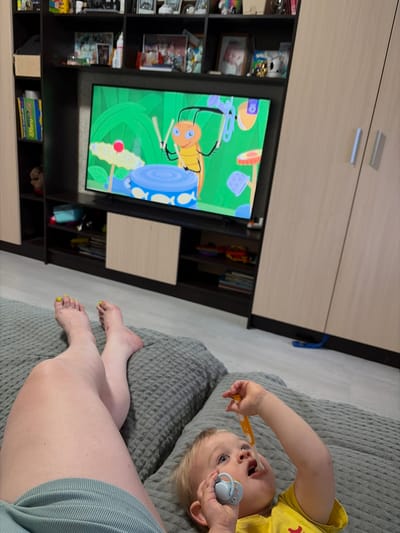 In a room with wooden floorboards and built-in dark brown shelving units, a television displays a cartoon with an orange insect character playing drums. To the right of the television, a light-colored wooden wardrobe with two doors and silver handles is visible. In the foreground, a person's legs and feet, with yellow nail polish, are resting on a grey textured surface. A baby, wearing a yellow shirt, lies on the same surface to the right, holding a pacifier and a yellow object. The environment suggests a residential setting in the city of Central District, Russia.