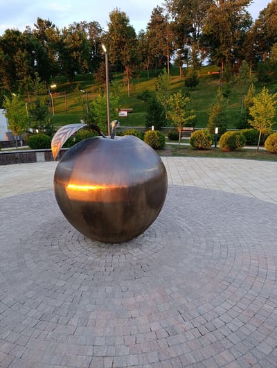 This is a medium shot, eye-level, outdoor photograph of a large, metallic apple sculpture situated in a public park. The sculpture is the primary subject, dominating the foreground and the left side of the frame. It appears to be made of a dark, polished metal, reflecting the warm, golden light of what seems to be either sunrise or sunset. A single, large, metallic leaf is attached to the top of the apple, also catching the light.
The apple is positioned on a paved circular area composed of small, gray cobblestones arranged in concentric rings. This circular plaza is bordered by a lighter, cream-colored paved area, which in turn gives way to a manicured grassy slope dotted with trees and shrubs.
In the background, a gently sloping hill is covered in lush green grass. Several trees of varying sizes and types are scattered across the hillside, with their leaves appearing either dark green or tinged with the warm light. There are several park benches visible on the slope, as well as lampposts providing illumination. The sky above is a pale blue with some wispy clouds, suggesting it is either early morning or late evening.
The scene appears to be a well-maintained urban park, likely a popular spot for leisure and recreation. There are no people visible in the image. The overall impression is one of peacefulness and natural beauty enhanced by artistic installation. The location is confirmed as the park on Lenin Street in the city of Klyuch, Russia.