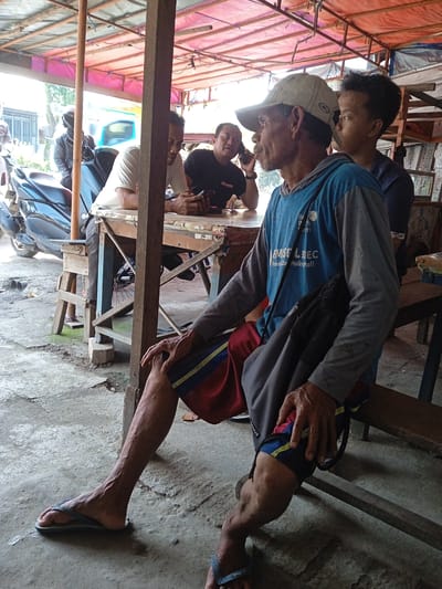 The image captures a candid scene in Sekopek Wetan, Indonesia. Four men are present under a corrugated metal roof structure. One man is seated on a wooden bench in the foreground, wearing a cap, long-sleeved blue shirt, red and blue shorts, and flip-flops. He has a black bag slung over his shoulder. Three men are seated at a wooden table; one is on a cell phone. A blue motorcycle is parked nearby. The ground is concrete, and the overall environment appears to be a roadside eatery or gathering place.