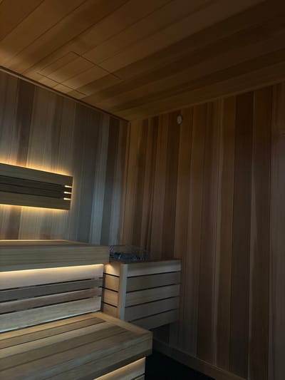 The image shows the interior of a wooden sauna, likely in Boca Raton, United States of America. The sauna is constructed with light-colored wooden planks on the walls and ceiling. Built-in wooden benches are visible in the foreground, with integrated LED lighting illuminating the seating areas. A section of the wall features a horizontal wooden accent with backlit lighting. To the right of the benches, a small wooden enclosure with horizontal slats houses a sauna heater with stones. The ceiling also appears to be constructed of wooden planks and has a recessed section. The overall lighting is warm and dim, contributing to a relaxing ambiance. There are no people or animals present in the image, and no visible action or interaction is occurring.
