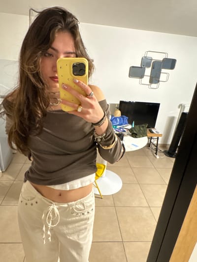 A young woman takes a mirror selfie in an interior space. She is wearing a brown long-sleeved shirt and white pants. The background includes a wall decoration consisting of geometric shapes, a television, and various objects on a shelf or counter. The floor is tiled. The location is not identifiable as Saint-Ouen-sur-Seine, France from this image.