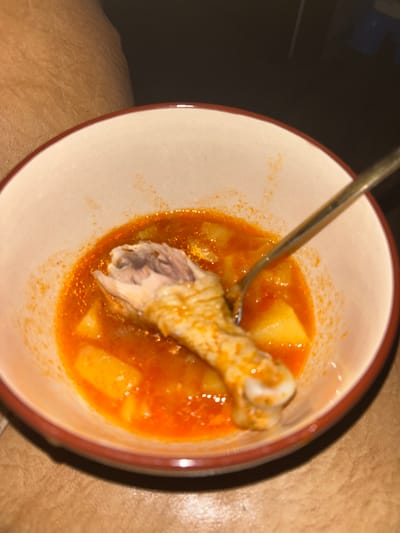 A close-up, overhead shot shows a bowl of food containing chicken, potatoes, and a reddish-orange broth. A fork is resting in the bowl, positioned over the chicken leg. The food is in a bowl with a light-colored interior and a reddish-brown rim. The background is dark and out of focus, with a hint of a textured surface that appears to be skin or fabric. There is no visible indication of the city of Dovatorovka, Russia, or any forensic evidence.