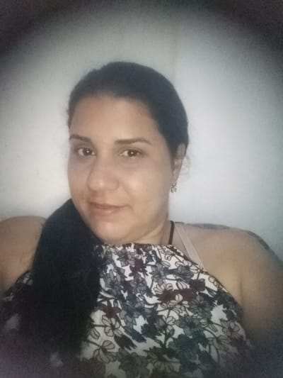 A woman is photographed in a brightly lit indoor setting against a white or light-colored wall. She has dark hair pulled back in a ponytail and is wearing a floral print top with a white strap visible underneath. She has earrings and a dark necklace. A tattoo is partially visible on her shoulder. The location is unknown.