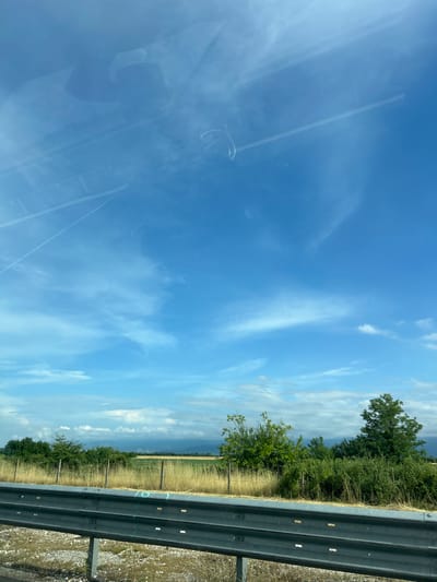 The image, captured from the interior of a vehicle, displays a bright, sunny roadside landscape, likely in the area of Malo Konare, Bulgaria. A grey metal crash barrier runs along the lower right side of the frame, indicating the edge of a road. Beyond the barrier, a strip of dry, golden-brown grass leads to dense green trees and bushes. Further in the background, agricultural fields, possibly of a cereal crop, extend towards hazy, distant hills or mountains.
The upper two-thirds of the image are dominated by a clear blue sky, adorned with scattered white clouds, ranging from wispy cirrus streaks to more defined cumulus formations. Reflections on the vehicle's window are noticeable on the left side, showing subtle outlines of the car's interior or perhaps a person, along with faint linear reflections that could be overhead power lines or contrails.
The scene depicts a daytime view under fair weather conditions, suggesting travel through a rural setting. On the crash barrier, a light blue spray-painted marking clearly shows the number "10", or possibly "L0", suggesting some form of road survey or maintenance identification.