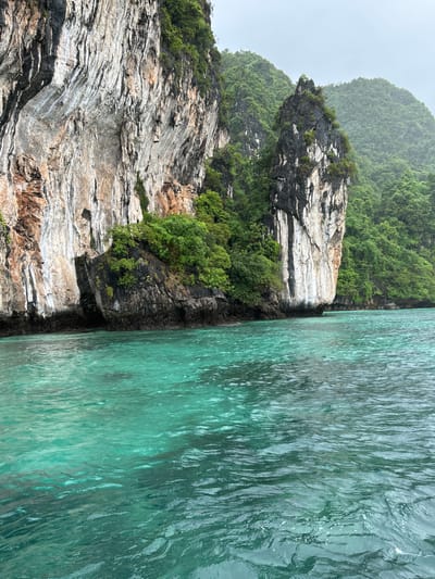 The image shows a view of the ocean near Haad Hin Khom, Thailand. A large, sheer limestone cliff face with visible strata and patches of green vegetation dominates the left side of the frame. To the right of this cliff, a smaller, darker, and more jagged rock formation juts out from the water, also covered in dense green foliage. The background is filled with lush, rolling hills covered in trees, suggesting a tropical environment. The water in the foreground is a clear, vibrant turquoise with gentle ripples, indicating calm seas. The sky is overcast and a pale grey, hinting at a cloudy or potentially rainy day. No people or man-made structures are visible in the scene, focusing entirely on the natural landscape.