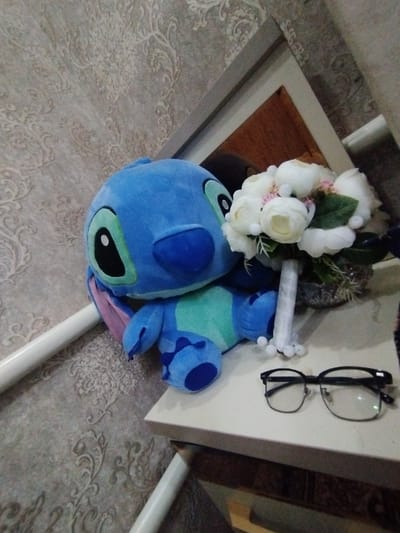A plush, blue Stitch toy sits next to a white floral bouquet on a table. Black-rimmed glasses rest beside the bouquet. The table is white and beige, and a white support beam is visible beneath it. The background features patterned wallpaper and a partially visible picture frame. The setting appears indoors, possibly a bedroom or living area. The lighting is soft and diffused.
