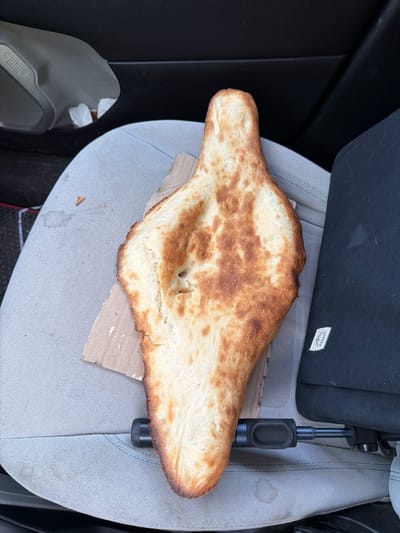The image shows a piece of flatbread resting on a car seat. The bread is light in color with golden-brown spots. It sits on a small piece of cardboard. The car seat is gray with blue stitching. A black object, possibly a car mount or phone holder, is adjacent to the bread. The brand "Osprey" is visible on a black bag behind the bread. A gray object with a Jeep logo can be seen in the lower-left corner. The location is not identifiable from the image.