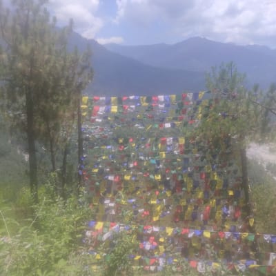 This is a photograph taken from a high vantage point overlooking the town of Baragran, India. In the foreground, a string of colorful prayer flags, a common sight in Buddhist regions, is draped across the frame, obscuring a portion of the view. Beyond the flags, the town of Baragran is visible nestled within a lush, green valley. Buildings with red and grey roofs are spread across the landscape. In the background, rolling mountains rise, fading into a hazy, blue-grey sky with scattered clouds. The overall impression is one of a serene, mountainous landscape with a touch of cultural vibrancy from the prayer flags.