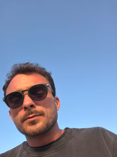 A male individual is depicted in a low-angle, close-up outdoor portrait, set against a clear, bright blue sky in Beit Yanai, Israel. The subject has short, dark brown, wavy hair, and a trimmed beard and mustache. He is wearing dark-framed sunglasses with light-colored accents on the upper corners and a dark gray crew-neck sweatshirt. A white earbud is visible in his right ear. Direct sunlight illuminates his face from the left, casting subtle shadows and highlighting his skin tone. The man is looking directly into the camera.