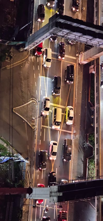 The image shows an aerial view of a busy intersection in Makati, Philippines at night. The scene is illuminated by streetlights and the headlights and taillights of numerous vehicles. The activity taking place is traffic, with cars, vans, and possibly a motorcycle moving through the intersection and on the surrounding roads. The roads are wet, suggesting recent rain. The overall mood is one of urban dynamism, with the constant flow of traffic indicating a typical evening in a bustling city.

The subjects are primarily vehicles of various types, including black SUVs, a yellow sedan, white vans, and a red car. The presence of multiple lanes and traffic signals indicates a major road junction. Overhead structures, likely part of an elevated road or bridge, are prominent in the frame, casting shadows on the road below. There is no visible text in the image. The time of day is clearly night, given the artificial lighting and the absence of daylight. The location context of Makati, Philippines, implies a densely populated urban environment with significant vehicular activity. The wet roads might also suggest a tropical climate, common in the Philippines. The composition of the image, with its high vantage point, emphasizes the scale and complexity of the traffic flow.