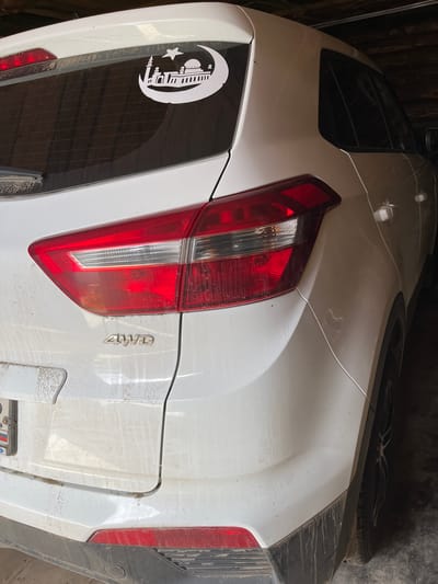 A white SUV parked indoors, possibly in a garage or storage area. The vehicle is covered in a layer of dust, with streaks visible on the rear door and bumper. A white sticker depicting a crescent moon with a mosque and a star is adhered to the rear window. The "4WD" badge is visible on the lower rear of the vehicle. The tail lights are red and clear. A license plate with Russian characters is partially visible on the left side of the rear. The surroundings are dimly lit and appear to be a rustic or industrial setting with wooden beams and dark walls. There are no people visible in the image.