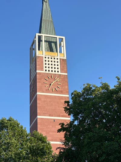 The image shows the tower of the Church of the Assumption in Pforzheim, Germany, on a clear, sunny day. The tower is made of red brick and features two large clocks. The top of the tower is a green, metallic spire. Lush green trees frame the bottom and right side of the tower. The sky is a vibrant blue, providing a stark contrast to the architecture and foliage. There are no people visible in the image.