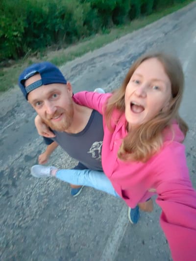 A man wearing a blue baseball cap and a dark t-shirt is giving a woman in a pink hoodie a piggyback ride. The woman has her mouth open and appears to be laughing. They are on a paved surface that looks like a road, with greenery on the left side and the road continuing into the background. The photo is taken from a high angle, looking down at the couple.