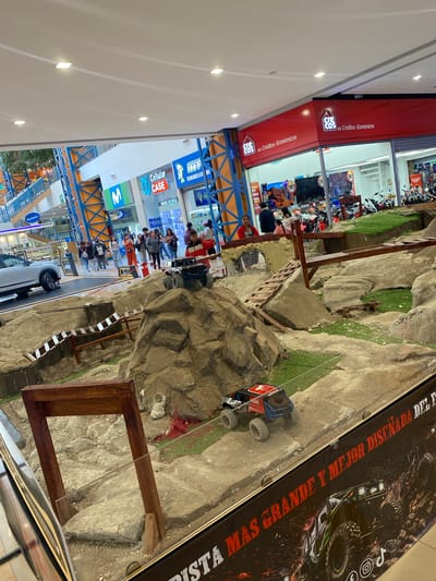 This image depicts the interior of a multi-level shopping mall in Quito, Ecuador, identified primarily by the prominent "Crecos es Créditos Económicos" retail store sign on the right.
In the foreground and extending into the midground, a large, elaborate miniature off-road course is displayed behind a transparent barrier. This course is constructed with faux rock formations, dirt-colored paths, and small patches of artificial green turf, simulating a rugged natural landscape. Two remote-control (RC) rock crawler vehicles are positioned on the track: a black RC vehicle with blue roll cage accents rests atop a central rock peak, and a red and blue RC vehicle marked with "2069" is situated on a lower dirt path. The course incorporates multiple wooden bridge structures and ramps, one of which forms a distinctive archway. A black banner along the bottom edge of the enclosure features red text, partially visible, that reads "PISTA MÁS GRANDE Y MEJOR DISEÑADA DEL E..." (Largest and best designed track of the...).
In the midground, approximately 15-20 individuals are present, walking, standing, and observing the various displays. They are dressed in casual attire. To the left of the RC track, a sleek silver or white SUV is parked on a raised display platform.
The mall's architecture features prominent blue and orange structural girders, creating a striking framework. Multiple retail establishments are visible in the background: "Crecos es Créditos Económicos" with its large red signage, "Cellular CASE" with blue and green branding, a large blue "M" logo (likely Movistar), and "HAPP Celulares & C...". Escalators are discernible in the far left background. The ceiling is white with evenly spaced square recessed LED lights. The flooring appears to be light-colored, possibly polished concrete or tile. The overall scene is well-lit and represents a modern commercial interior.