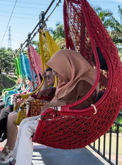 In the city of Bintang Meriah, Indonesia, a group of people are enjoying a ride on a series of colorful, netted hanging chairs. The chairs, which are red, yellow, pink, and blue, are suspended from a cable system. A woman wearing a light brown hijab and a brown shirt is seated in the foreground in a red chair, looking down. Behind her, several other individuals are visible in the other chairs. The background shows a lush green environment with palm trees, a body of water, and a transmission tower in the distance. The sky is clear and blue.