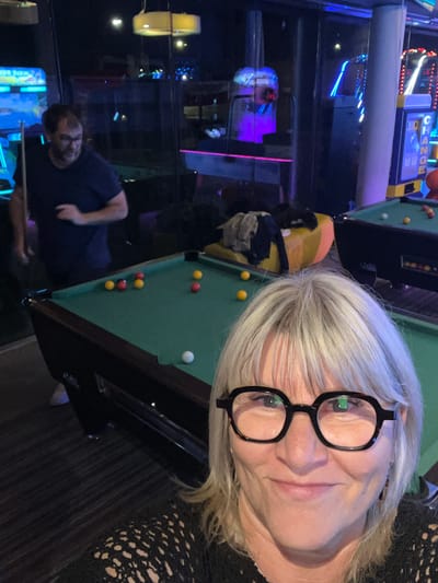 The image depicts a selfie of a woman with short, gray hair and black-framed glasses, taken indoors in what appears to be an arcade or amusement center. She is positioned near a pool table with several billiard balls scattered across the green felt. A man is visible in the background, holding a pool cue and seemingly preparing to take a shot. Various arcade games and illuminated displays are also discernible in the background, adding to the ambiance. Based on this information, the location is Montrond-les-Bains, France.