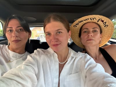 Three women are seated in the backseat of a car, taking a selfie. The woman on the left has dark hair and is wearing a white t-shirt and gold earrings. The woman in the middle has blonde hair pulled back and is wearing a white linen shirt and a pearl necklace. The woman on the right is wearing a straw hat with black lettering, a black top, and hoop earrings. The background shows a building with orange rooftops, suggesting an urban or suburban setting. The car interior is visible, with the car's ceiling and windows in the frame. The lighting suggests it is daytime. The location is the city of Šibenik, Croatia.