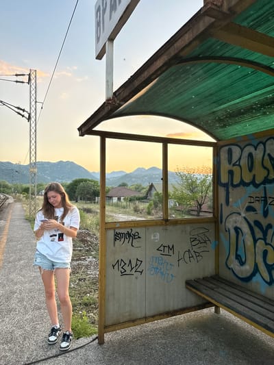 In Vranjina, Montenegro, at a small, rustic train station, a young woman stands on the platform, engrossed in her phone. She wears a white t-shirt and denim shorts, with black and white sneakers. The station features a covered waiting area with a green corrugated plastic roof and a wooden bench. The walls of the shelter are heavily adorned with graffiti, including tag-style writing in yellow, blue, and black. Beyond the station, the landscape opens up to rolling hills and mountains, illuminated by the warm glow of a sunset. Train tracks run alongside the platform, disappearing into the distance. The sky is a gradient of pale blue and soft orange, with a few wispy clouds. The scene captures a moment of quiet contemplation at a remote, somewhat weathered, but picturesque location.