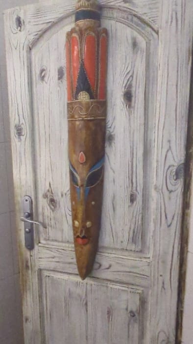 The image displays an elongated, decorative wooden mask mounted vertically on a light-colored wooden door. The mask is predominantly brown, featuring a stylized human face with an ornate headdress-like top. The upper section of the mask is painted with red, dark blue, and gold patterns, including a pointed, crown-like element. Mid-section details include stylized blue and black eyes with white outlines, a small red teardrop-shaped inlay on the forehead, and two small golden circular elements on the cheeks. The lower section tapers to a point, featuring a red mouth. The background is a paneled wooden door, distressed or white-washed, exhibiting prominent wood grain patterns and scattered dark circular knots. A silver-toned metallic door handle and escutcheon are visible on the left side of the door. This scene is located in Slokoshtitsa, Bulgaria.
