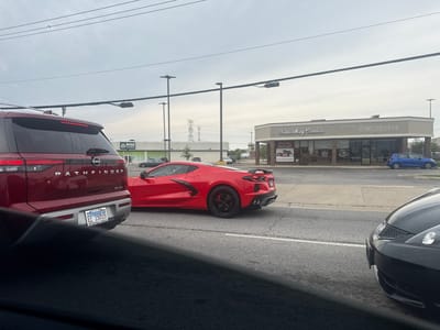 In Niles, Illinois, USA, a red Nissan Pathfinder SUV and a red Chevrolet Corvette are stopped on a road, likely at a traffic light, with other vehicles around. The Pathfinder, in the foreground on the left, has its rear visible, showing a license plate with the text "EL 15055" and "WOODFIELD NISSAN." To the right of the Pathfinder, the red Corvette is positioned, showcasing its sleek design and black wheels.
In the background, a strip mall is visible, featuring businesses such as "Fannie May Candies," an "Outlet," and another establishment with the word "MOCHINUT" visible on its signage. A blue compact car is parked in front of one of the stores. Power lines and utility poles stretch across the sky, and distant industrial structures are visible. The sky is overcast.