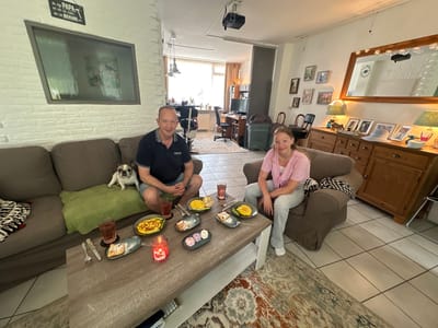 In Steenwijk, Netherlands, a man and a young girl are seated at a coffee table in a living room. The man, wearing a dark blue polo shirt and denim shorts, is on the left side of the frame, with a French bulldog resting on a green cushion beside him. The girl, in a pink top and light blue jeans, is on the right.
The coffee table is laden with food and drinks. Plates of what appears to be omelets are visible, along with a dessert with small pastel-colored mounds and a pastry. Two glasses of red liquid and a lit red candle are also on the table. Utensils, including forks and a knife, are arranged near the plates.
The living room features a dark brown sofa and armchair. In the background, a doorway leads to another room where a desk with a computer and a projector mounted on the ceiling are visible. The walls are painted white, and decorative items such as framed pictures and a large mirror are displayed. A patterned rug covers a portion of the tiled floor.