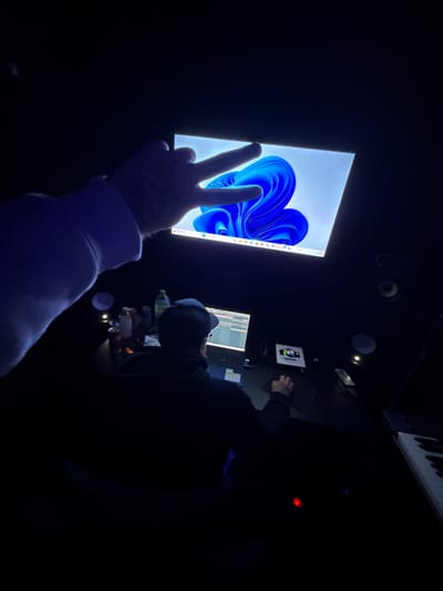 This image captures a dimly lit indoor studio scene, situated in Kassel, Germany. In the upper left foreground, a left hand, clad in a light-colored long-sleeved garment, extends into the frame, forming a two-finger "peace" or "V" sign. A tattoo is faintly visible on the back of this hand. Below and centrally positioned, a second individual is seated with their back oriented towards the camera, facing a dark desk. This person wears a dark hooded top and a dark baseball cap, and is actively using a laptop computer, which displays what appears to be a Digital Audio Workstation (DAW) interface. Their right hand operates a computer mouse on the desk.
Above the desk, a large, brightly illuminated monitor prominently displays the standard blue, flowing abstract wallpaper of the Windows 11 operating system. The taskbar with various icons is visible at the bottom of this screen. The desk itself is dark and holds several objects: a clear plastic water bottle, a dark bottle, a red Coca-Cola can, a white-and-silver can, and other small containers to the left of the laptop. To the right of the laptop, a white square device with a central glowing green circular element is present, likely an audio interface or controller. Studio monitor speakers are partially visible to the upper right of the main monitor and also on the desk to the right. A section of a black and white electronic musical keyboard is visible in the lower right corner of the frame. Illumination in the scene primarily stems from the computer screens and small, discrete light sources on the desk.