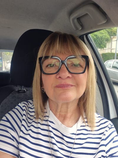 A medium shot captures a woman from the chest up, seated inside a car. She has blonde hair with bangs and wears large, black-rimmed glasses. A blue and white striped t-shirt adorns her, with a silver necklace visible. She has a subtle smile and wears small hoop earrings. The background shows the car's interior, including a headrest and the ceiling handle. Outside the car, trees and buildings are visible, suggesting an urban setting. The car's side window is open. The image's context suggests Póvoa de Varzim, Portugal.