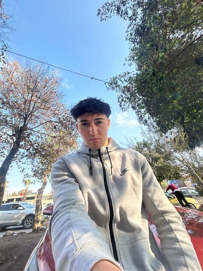 A daytime image taken in Al-Qaboun Municipality, Syria, depicts a young male in the foreground. He possesses dark, curly hair and maintains a neutral expression while looking directly at the camera. He is wearing a light grey full-zip hooded sweatshirt featuring a black zipper and a black Nike swoosh logo on the left chest. His right arm is visible, extended towards the camera, consistent with a self-portrait. He appears to be leaning over or positioned in front of a partially visible reddish-orange surface, likely the hood of a vehicle.

The background reveals a clear blue sky with scattered wispy clouds. To the left, a deciduous tree with sparse brown leaves is visible. Further back, a white SUV is parked on a street, with indistinct buildings in the distance. To the right, a tree with dense green foliage is present. Below this tree, a second individual, wearing a dark red long-sleeved shirt and dark pants, is seen leaning over the hood of a yellow vehicle. A thin dark utility wire spans across the upper-mid portion of the frame against the sky.