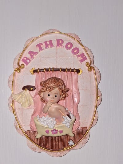 A decorative plaque reads "BATHROOM" in pink lettering against a light pink background. The plaque depicts a young child sitting in a bathtub filled with white bubbles. The child has curly brown hair and is reaching a hand towards their mouth. Pink curtains hang behind the bathtub, and a small yellow towel is draped over a hook on the left side of the plaque. The plaque is framed with a scalloped pink edge and gold trim. The background appears to be a white wall. The image is a close-up of the plaque, and no specific location in Åsgårdstrand, Norway, is visible.