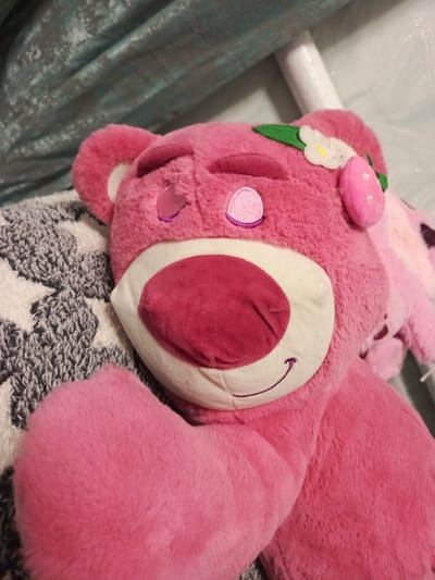 A close-up, slightly angled shot shows a plush toy bear, Lotso Huggin' Bear from Toy Story, lying down. The bear is a bright pink color and appears to be made of soft, fuzzy material. It has embroidered features including closed eyes with purple stitching, a light pink nose, and a smiling mouth with purple stitching. A small, white felt flower with a yellow center and a green leaf is attached to the top of its head, along with a pink strawberry-shaped accessory. The bear is resting on a gray and white patterned blanket that features star shapes. In the background, a draped teal-colored fabric can be seen. The image is well-lit, with some reflections visible on the background fabric. There is no indication of the location being in Вылково, Russia.