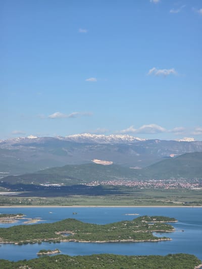 The image shows a landscape with a clear blue sky dotted with a few wispy clouds on the left. Moving right, snow-capped mountains transition into green hills and a cityscape. A narrow strip of green land separates the town from a large body of blue water. Several small, tree-covered islands are scattered in the lake, appearing parallel to the mountains, along the city of Ponikvica, Montenegro.