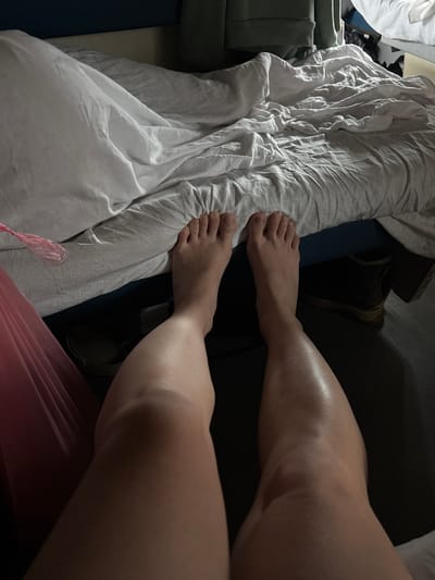 A first-person perspective in an indoor setting in Kargat, Russia, captures the bare legs and feet of an individual, extended forward. The person is positioned on a bed covered with wrinkled white sheets or a duvet. A small, crinkled pink translucent plastic object is visible on the left side of the bed, near the left leg. Under the bed, on the right, a pair of dark-colored boots or similar footwear is partially visible on the floor. The background features a dark blue surface, likely a headboard or wall, and a crumpled green garment or object resting on the bed's edge further back. The scene is illuminated by dim, ambient light.