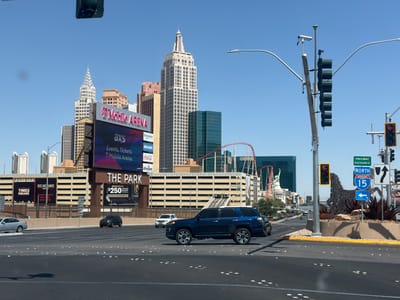The image depicts a street view in Paradise, Nevada, USA. In the background, several iconic Las Vegas buildings are visible, including replicas of the Chrysler Building and the Empire State Building. A large billboard promoting T-Mobile Arena and "The Park" is prominent, advertising events and betting offers.
In the foreground, a blue SUV is crossing an intersection. Street signs indicate the entrance to Freeway I-15 North. A decorative sculpture of a horse is present on the median. Traffic signals and streetlights are visible. Other cars can be seen on the road. The weather appears to be sunny.