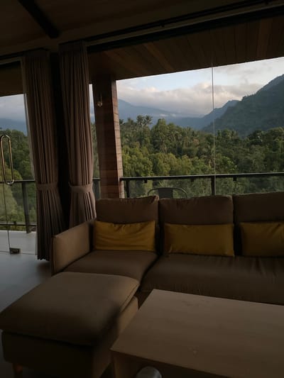 A low-angle, vertical shot captures an interior scene with a view of lush, green mountains and cloudy skies. The room features a large, sectional sofa with several cushions, some in a muted brown and others in a bright yellow. To the left of the sofa, a wall with wooden paneling and a window with drawn brown curtains are visible. Through the window, the dense foliage of trees and the layered slopes of mountains are seen under a bright, overcast sky. The scene appears to be from a balcony or a room with a large window overlooking a natural landscape. The lighting suggests it is daytime, possibly late afternoon or early morning due to the soft light. There is no visible text in the image. The setting is likely a hotel room or a private residence designed to offer scenic views, possibly in a tropical or mountainous region like Sawan, Indonesia, given the dense greenery. The overall mood is one of tranquility and relaxation, with the comfortable seating inviting one to enjoy the natural panorama.