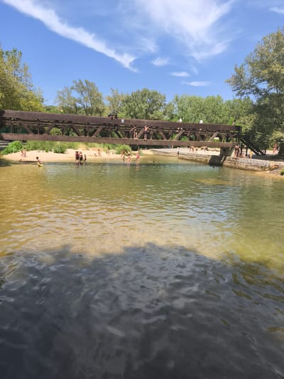 The image depicts a sunny day with a clear blue sky and scattered white clouds. A wooden bridge spans across a body of water, which appears to be a river or a wide stream. The water is shallow in some areas, revealing sandy or rocky bottoms, while other parts are deeper and appear darker. Several people, mostly children and young adults, are visible in and around the water. Some are wading, others are standing on the sandy bank, and a few are on the bridge, with some appearing to be preparing to jump or having just jumped into the water. The bridge is constructed of dark wood with visible support beams and railings. Trees and lush green foliage line the banks of the water, suggesting a natural, outdoor setting. The overall scene conveys a sense of recreation and enjoyment on a warm day. The specific location within Campezo / Kanpezu, Spain, is not discernible from the visual information provided in the image.