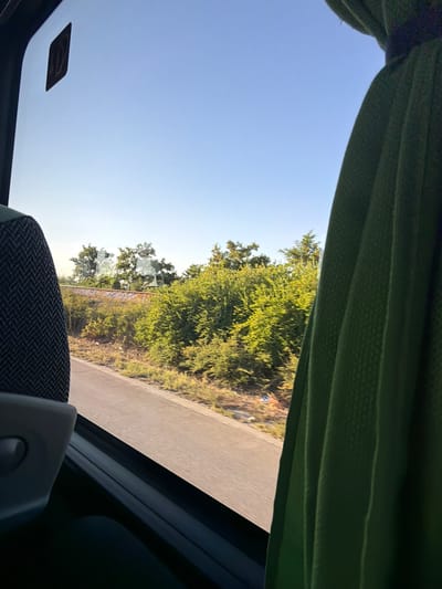 The image shows a view from inside a vehicle, likely a bus, looking out the window. The left side of the frame is occupied by the back of a seat, upholstered in a dark fabric with a patterned weave. To the right, a green curtain hangs, partially obscuring the view.
Through the window, a road runs parallel to a stretch of greenery dominated by lush trees and bushes. In the distance, beyond the vegetation, a railway track can be seen. The sky above is a clear, bright blue, suggesting a sunny day. A small, square sticker with an upward-pointing arrow is visible on the window in the upper left corner. The overall impression is of a journey through a rural or semi-rural area near Benkovski, Bulgaria, possibly on a highway adjacent to a railway line.