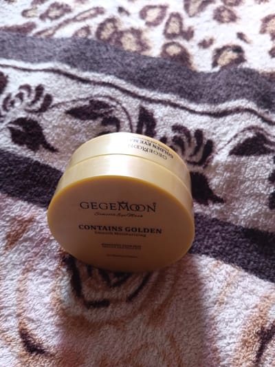 A circular, golden jar of eye masks is positioned on a textured, brown and white patterned cloth. The jar has text printed on it, with the brand name "GEGEMOON" prominently displayed in black lettering, followed by "Smooth Eye Mask". Below this, it states "CONTAINS GOLDEN" and "Smooth Moisturizing". The jar is slightly angled, with the lid slightly askew, revealing a portion of the contents. The cloth beneath the jar appears to be a blanket or rug with a repeating leopard-like print. The lighting suggests an indoor setting, with shadows cast by the jar. There are no people or animals visible in the image. The image does not provide any information about the location in the city of Monastirische, Russia.