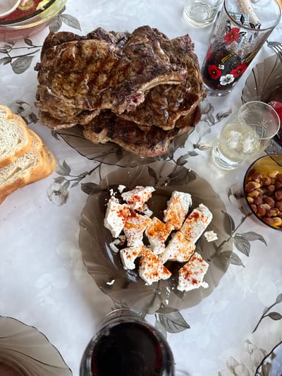 A close-up, overhead shot captures a feast laid out on a white tablecloth with a floral pattern, set on a table at an outdoor location, possibly a restaurant or a home courtyard, in Ravadinovo, Bulgaria. The spread includes several grilled steaks with a dark sear, piled on a dark grey, wavy-edged plate. To the left of the steaks, a stack of sliced white bread rests on the tablecloth. In the foreground, another dark grey plate holds pieces of white cheese sprinkled with red spice, likely paprika. A small, amber-colored bowl to the right contains roasted nuts. Various glasses are scattered across the table: one tall, clear glass with ice, another tall glass decorated with red and white flowers containing a dark liquid, and a third glass, partially visible, also with a dark liquid. The scene suggests a leisurely meal, possibly lunch or dinner, enjoyed in a relaxed outdoor setting characteristic of Ravadinovo, a village known for its fairytale castle and seaside charm, though the specific location within the village is not identifiable from the image alone.