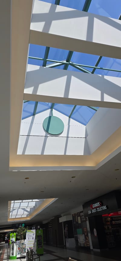 The image depicts the interior of a modern shopping mall with a prominent skylight. The skylight, constructed with glass panels and a metal frame, allows natural light to illuminate the space, casting geometric shadows on the white ceiling and walls. A large, circular, teal-colored decoration is visible on the upper part of the wall beneath the skylight. The lower portion of the image shows a mall concourse with various shops. One shop, labeled "GAMES" and "JEUX - JOUETS - COLLECTIONS," is clearly visible on the right. To the left, other retail displays and entrances are present, along with seating areas. The overall environment suggests a well-lit and contemporary commercial space. This mall is identified as being located in Victoriaville, Canada.