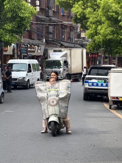 A person on a scooter travels down a street lined with parked delivery trucks, in the town of Fenghuang, China. The person on the scooter is seen through a plastic rain shield with a bear pattern. Buildings, trees, and a Luckin Coffee sign are visible. The person's destination is unknown.