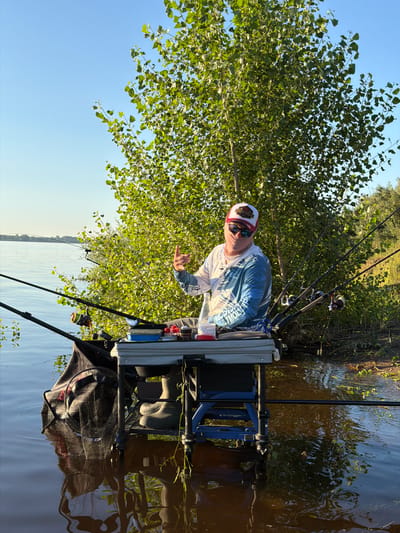 A man wearing a fishing hat, sunglasses, and a long-sleeved shirt with a blue and white pattern is fishing on a calm body of water. He is sitting behind a fishing seat with several fishing rods propped up. The man is holding up his thumb in a gesture of approval. A net bag is attached to the side of his fishing seat, and it is partially submerged in the water. The surrounding environment is lush with greenery, with a large tree providing shade. The water reflects the sky and the surrounding vegetation, creating a serene atmosphere. The location appears to be a natural setting, possibly a lake or riverbank, near Tarasovo, Russia.