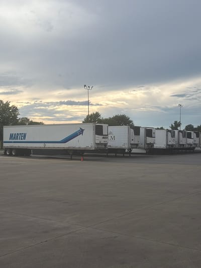 The image shows a line of refrigerated semi-trailer trucks parked in an asphalt lot. The trucks are white with varying logos and markings. One truck prominently displays "MARTEN" in large blue letters with a blue arrow graphic. Another truck has a logo with "LM SERVICES". The sky is overcast with some clouds, and there are trees visible in the background. A tall lamp post with multiple lights stands in the middle ground. The scene appears to be in a commercial or industrial area, possibly a truck depot or distribution center. The location is identified as Murfreesboro, United States of America.
