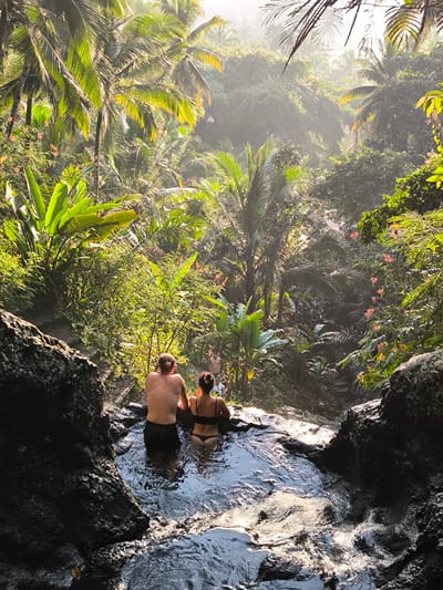The photograph, taken in the city of Sidemen, Indonesia, depicts a couple sitting in a natural pool situated at the edge of a waterfall, surrounded by lush tropical vegetation. The man, shirtless and facing away from the camera, is wearing dark-colored shorts. The woman, also facing away, is wearing a dark bikini. The pool is formed by dark, rugged rocks. Water flows over the edge, creating the waterfall. The scene is enveloped by dense foliage, including palm trees and various plants, indicative of a tropical environment. Other people are visible further into the background. Sunlight filters through the dense canopy, creating bright spots.