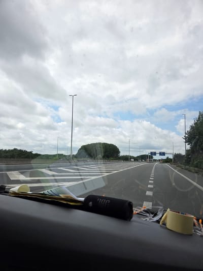 The image shows a view from inside a vehicle driving on a highway. The perspective is angled, with the highway and the sky dominating the frame. The highway is paved and has clear white lane markings, including a solid white line and dashed white lines. On the right side of the highway, there are several blue road signs with white text indicating directions to cities like Gent and Kortrijk, and an arrow pointing to "A17". There are also red and white directional signs.
Along the sides of the highway, there are tall lamp posts and lush green trees. The sky is filled with a mix of white and grey clouds, with patches of blue sky visible, suggesting an overcast or partly cloudy day.
Inside the vehicle, on the dashboard or a similar surface in the foreground, are several objects. A black cylindrical object, possibly a speaker, with "JBL" visible on it, is secured with tan tape. Some papers and a white disposable cup are also visible.
The overall scene is a typical highway drive on a cloudy day in Belgium, likely in the vicinity of Kortrijk, as indicated by the road signs. There are no people visible in the image. The time of day appears to be daytime, given the brightness and sky conditions. The mood is neutral, focusing on the travel scenery.