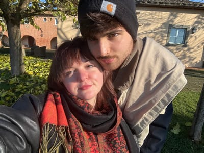 The image captures two young adults, a female and a male, in a close-up outdoor selfie. The female, positioned in the foreground and to the left, has brown hair with bangs, fair skin, and is wearing a black leather jacket and a red and black patterned scarf with fringe. Her face is angled slightly forward with a faint smile. The male, positioned behind and to the right of the female, has dark hair, a beard, and a nose ring on his left nostril. He wears a black Carhartt beanie with a visible Carhartt logo and a beige collared jacket over a dark garment. His expression is neutral.
The scene is set outdoors in Montepulciano, Italy. In the background to the left, a multi-story brick building with arched openings and a tiled roof is visible, suggesting traditional Tuscan architecture. Outdoor seating furniture is discernible beneath these arches. To the right background, a lighter-colored, possibly stucco, building features a single window and an exterior wall lantern. Long shadows are cast across this building, indicating strong, low-angle natural sunlight, suggestive of morning or late afternoon. The ground is covered with green grass and numerous fallen yellow-green leaves, indicative of autumn. A tree with sparse foliage is present in the mid-ground.