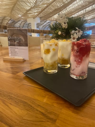 The image displays a close-up view of a table in what appears to be an indoor cafe or restaurant. On a dark tray, three glasses filled with colorful beverages are arranged. The glass on the right contains a pink liquid layered with red fruit pieces, likely a berry smoothie. The two glasses to its left contain lighter colored drinks with visible fruit chunks, possibly mango or peach. In the background, a membership card with the word "QUANTUM" and "PREMIUM MEMBERSHIP" is visible on a wooden stand. Behind the drinks, a small festive arrangement with pine branches, white flowers, and ornaments is set up. The ceiling is constructed with woven natural materials, and the table surface is made of polished wooden planks. The overall atmosphere suggests a comfortable and somewhat upscale dining or relaxation space. The image is tagged with the location "Gimpo-si, South Korea."