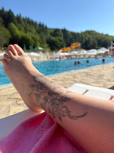 A person's foot and lower leg are shown in the foreground, with a floral tattoo visible on the leg. The person is lying on a white lounge chair with a pink towel draped over it. In the background, a swimming pool is filled with water, and several people are swimming. Beyond the pool, a lush green forest covers a hillside under a clear blue sky. Water slides and white umbrellas are also visible, suggesting a water park or resort setting. The scene appears to be located in the city of Chiflik, Bulgaria.