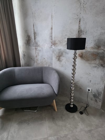 An indoor scene shows a grey upholstered loveseat with wooden legs and a floor lamp with a black shade and a silver beaded stand. The loveseat is positioned to the left, and the floor lamp is to the right of it, standing next to a wall with a textured, distressed tile finish. A brown curtain is visible on the far left. Underneath the loveseat, a silver digital scale is partially visible. The floor is tiled with light grey square tiles. A white electrical outlet with a black plug is on the wall to the right of the floor lamp. There are no people in the image. The setting appears to be a room in a residential or commercial space in Аснежыцкі сельскі Савет, Belarus.