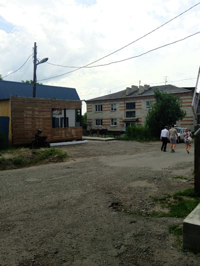 The image depicts a street scene in Pavlovsk, Russia. In the foreground, a gravel road leads towards two buildings. On the left, a modern-looking structure with horizontal wooden slats and a dark roof is prominent. A black motorcycle is parked in front of it. To the right, a multi-story brick building with orange accents and several windows stands under a cloudy sky. Three people are walking away from the camera on the gravel road, heading towards the brick building. One person is wearing a white shirt and dark pants, another a light-colored t-shirt and shorts, and the third is wearing a patterned dress. A white SUV is parked near the brick building. The scene appears to be set during the daytime, with overcast weather. Power lines are visible overhead, strung between utility poles. There is some greenery around the buildings and growing from the gravel road. A sign with text in Cyrillic is partially visible on the wooden structure.