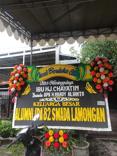 A large, elaborate funeral wreath (papan bunga duka cita) stands prominently, conveying condolences. The wreath features a black rectangular board adorned with vibrant yellow text and colorful floral arrangements at the top and bottom.
The text on the board, which is yellow against the black background, reads: "Turut Berduka Cita" (Deepest Condolences) at the top, followed by "Atas Meninggalnya" (For the passing of). The deceased is identified as "IBU HJ.CHAYATIN," who is further specified as "Ibunda BPK H.HANDY ALIANTO" (Mother of Mr. H. Handy Alianto). The senders of the condolence message are "KELUARGA BESAR" (Extended Family) and "ALUMNI '82 SMADA LAMONGAN," indicating a group of alumni from a high school in Lamongan from the graduating class of 1982. Decorative elements include yellow butterfly outlines and intricate patterns.
The floral arrangements feature dense clusters of red, orange, yellow, and white flowers, with hints of pink and purple blooms, creating a solemn yet colorful display.
The wreath is situated outdoors, possibly under a covered structure with a wavy-edged roof, visible in the background along with green foliage and a clear sky. A black motor scooter is parked in the foreground to the right, partially obscuring the bottom right portion of the wreath. A potted plant, possibly a cactus, can be seen behind the wreath in one of the crop images. The setting suggests a memorial event in Lamongan, Indonesia, during daytime.